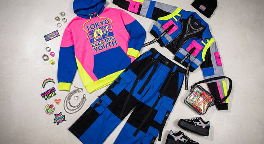 A flat lay editorial arrangement of a Harajuku streetwear outfit including an oversized graphic hoodie, wide-leg cargo pants, and a bold statement jacket surrounded by accessories on a concrete surface.